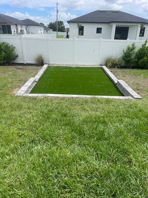 Green artificial turf inset in a rectangular concrete frame, surrounded by a grassy yard, a white fence, and houses.