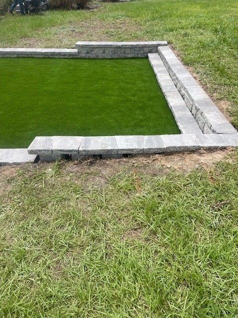 A rectangular area of green turf bordered by grey stone blocks and steps, set in a grassy yard.