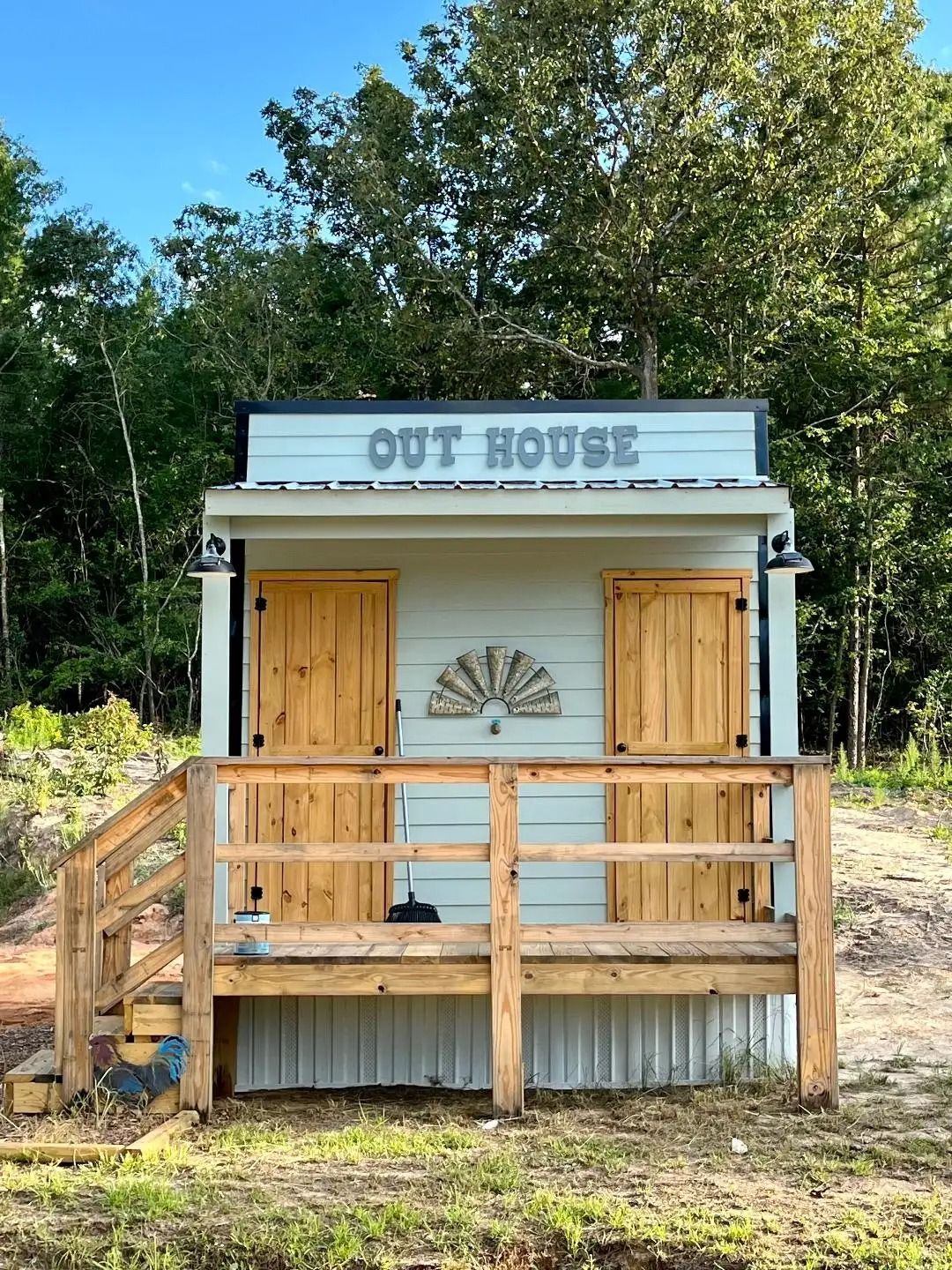 Small white outhouse with wooden porch and “OUT HOUSE” sign in a wooded yard.