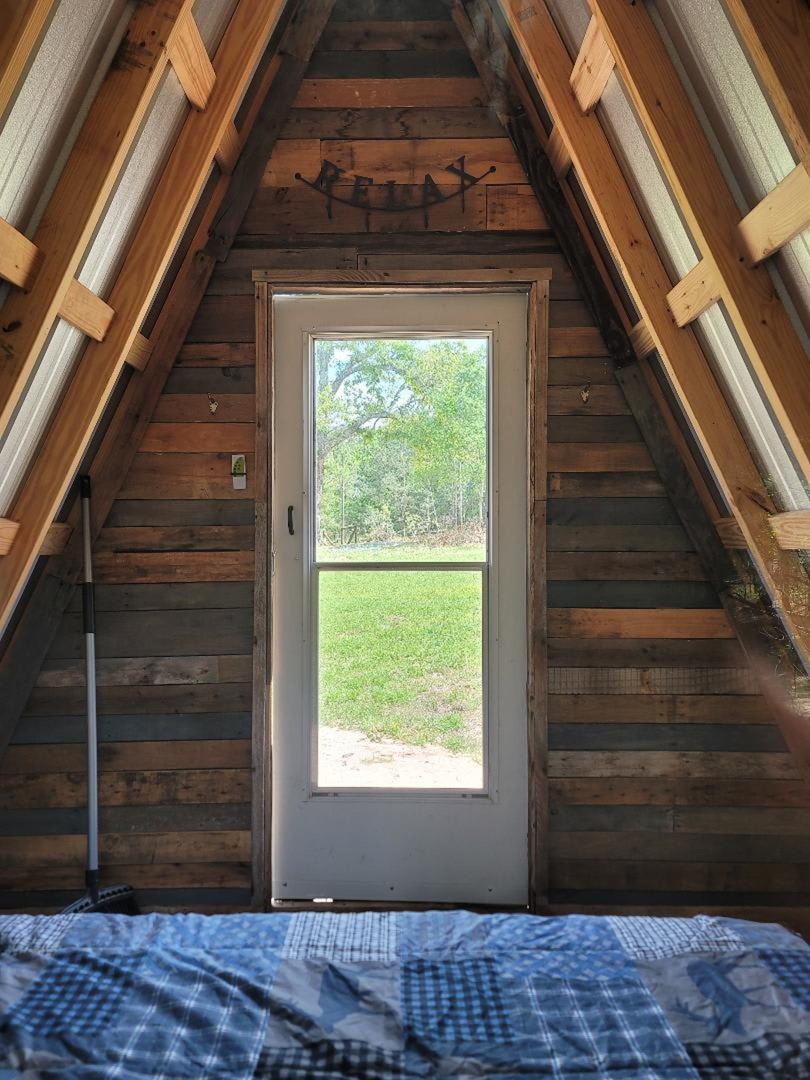 A-frame cabin interior with a narrow door centered on a grassy yard view and a blue bed in the foreground