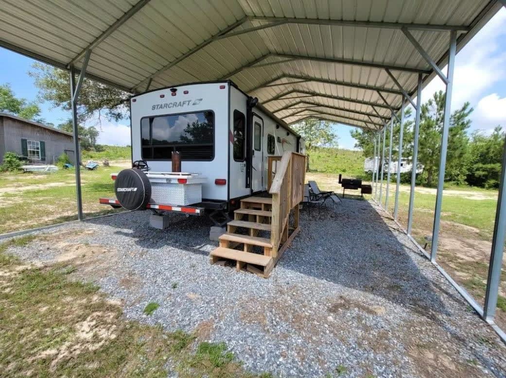 RV parked under metal carport with wooden steps and gravel ground