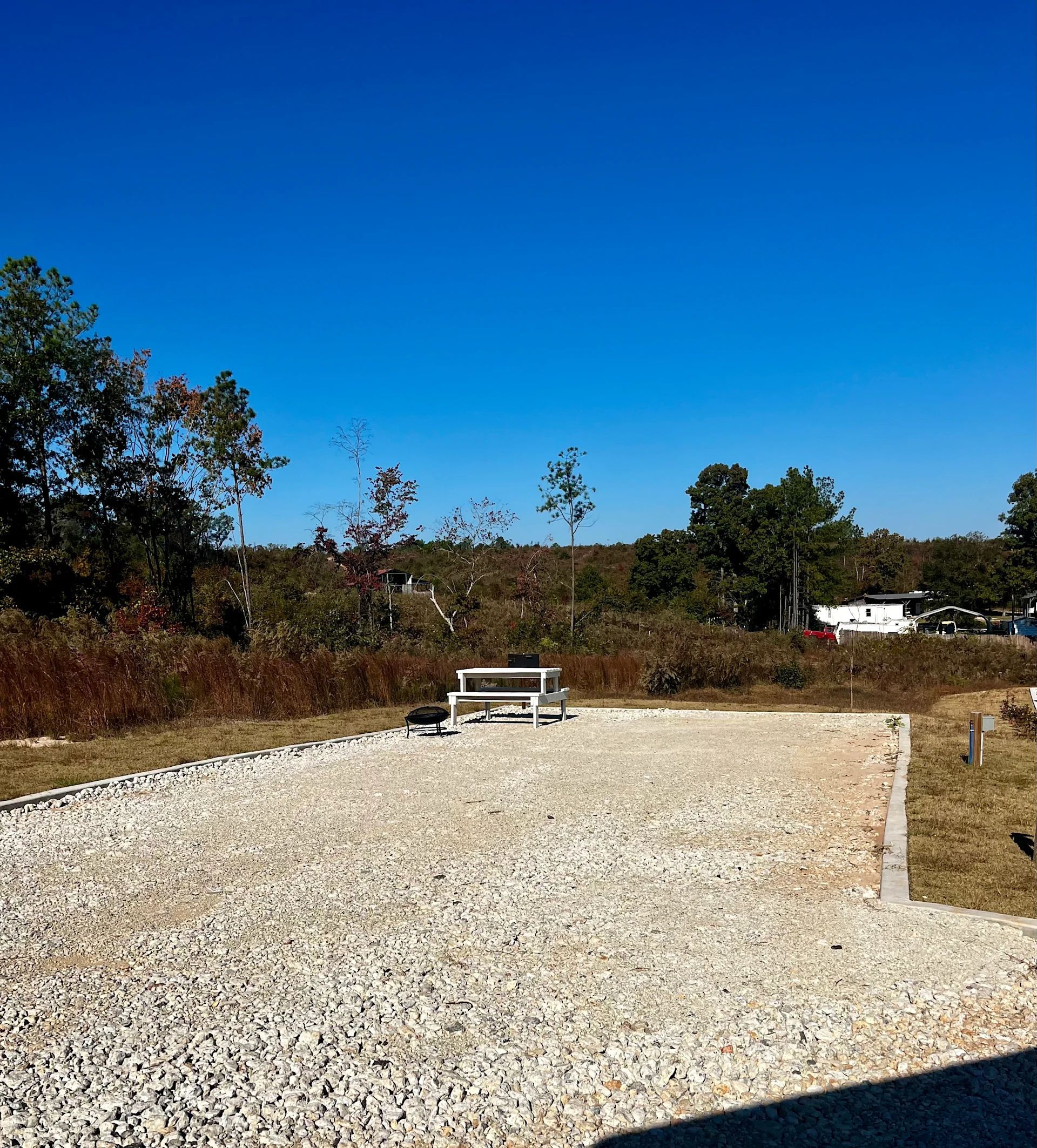 Gravel path with a white bench under a clear blue sky, bordered by dry grass and trees.