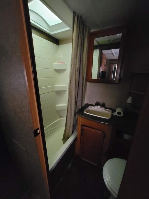 Narrow RV bathroom with shower, sink, toilet, and overhead skylight, in beige and wood tones