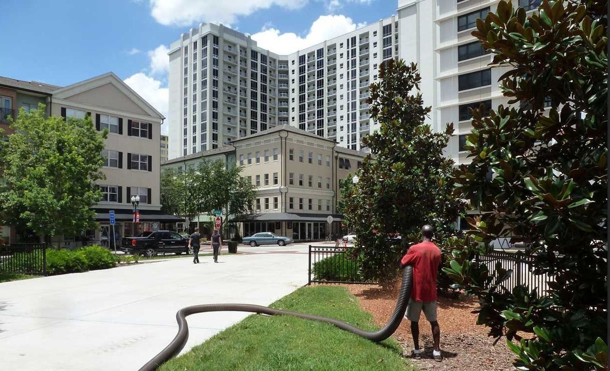 A man is using a vacuum hose to clean a lawn in front of tall buildings