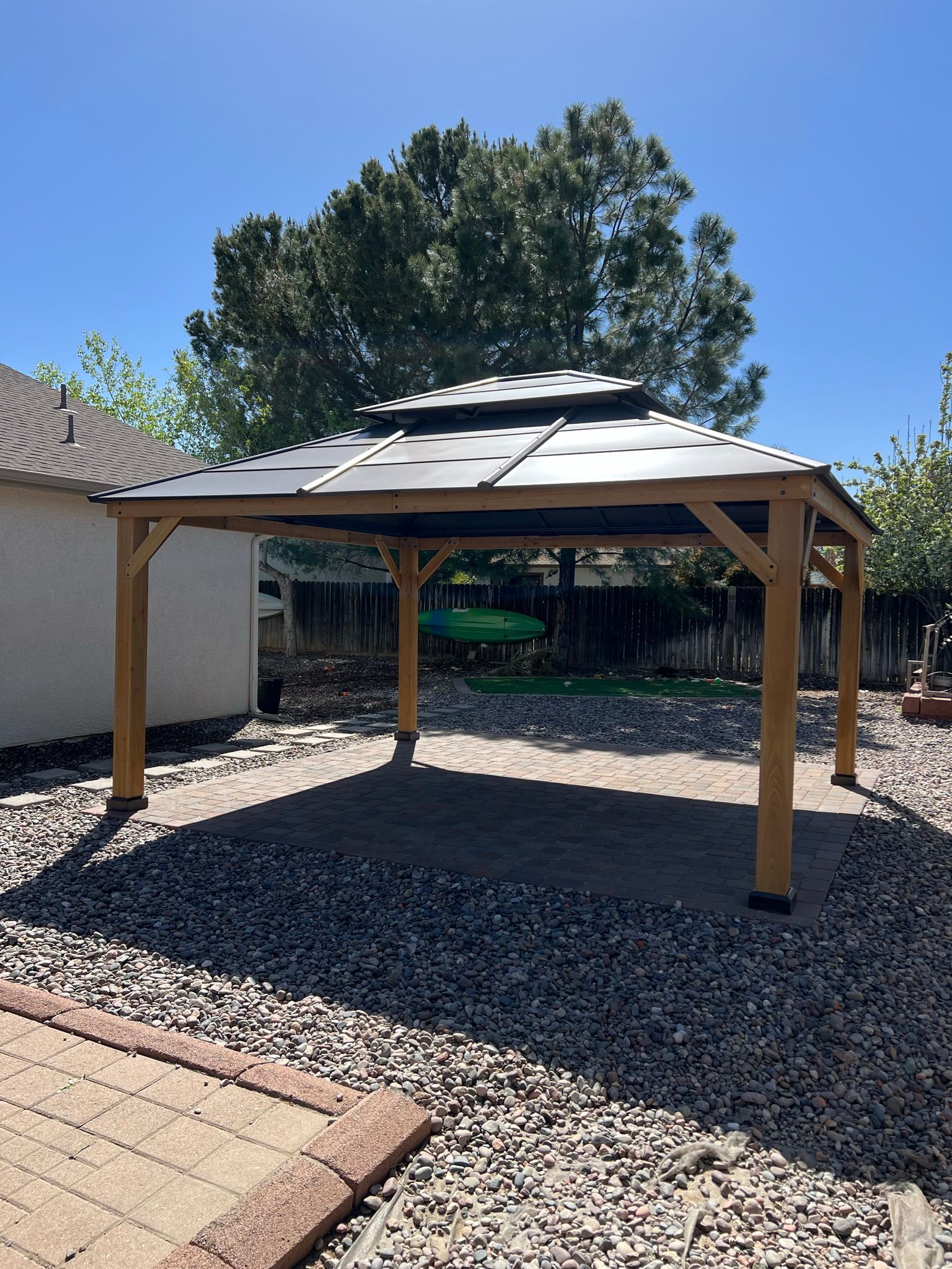 A wooden gazebo is sitting in the middle of a gravel yard in front of a house.