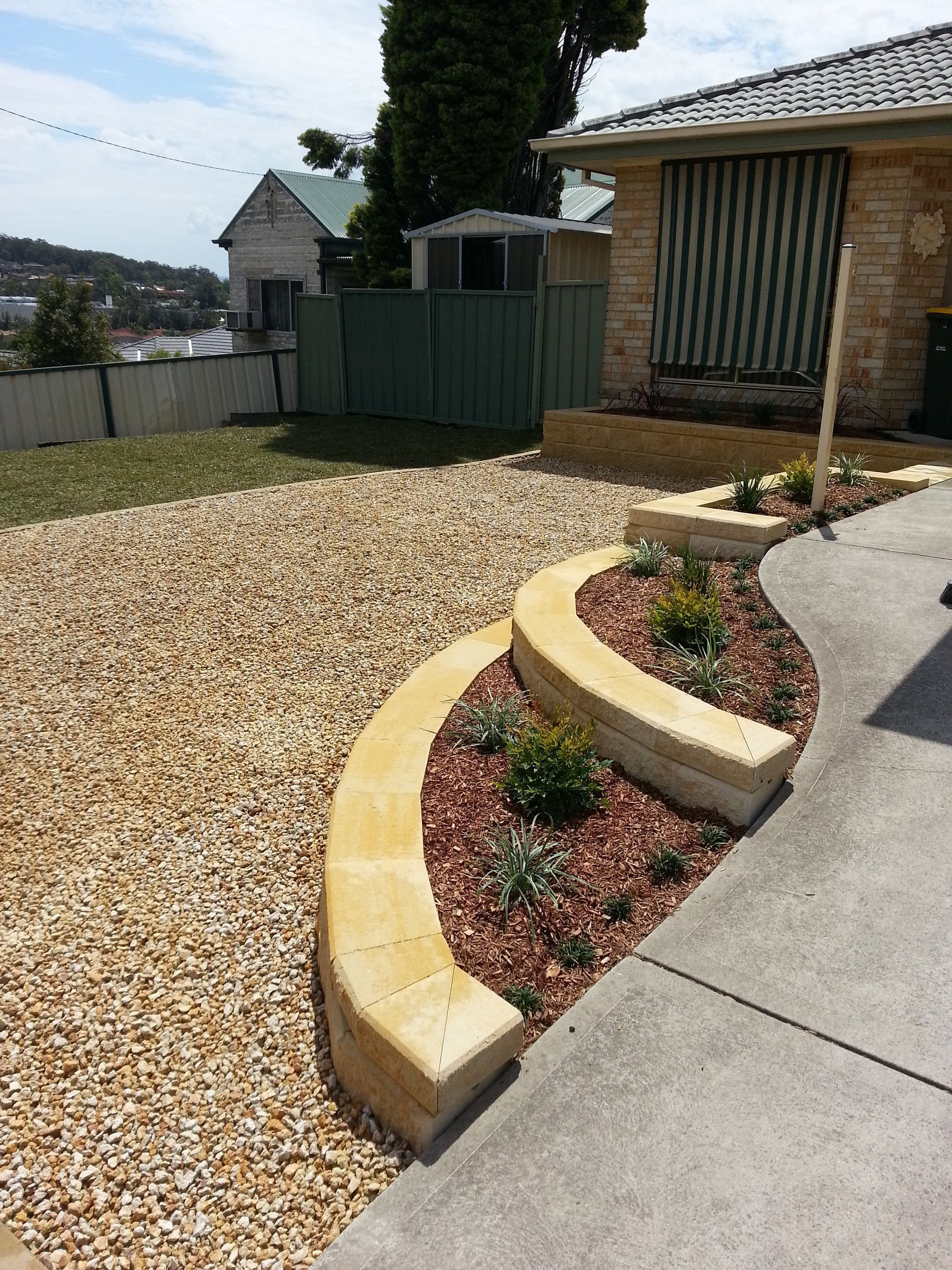 Curved, tan stone retaining wall with plants and gravel driveway next to a house with a concrete driveway.