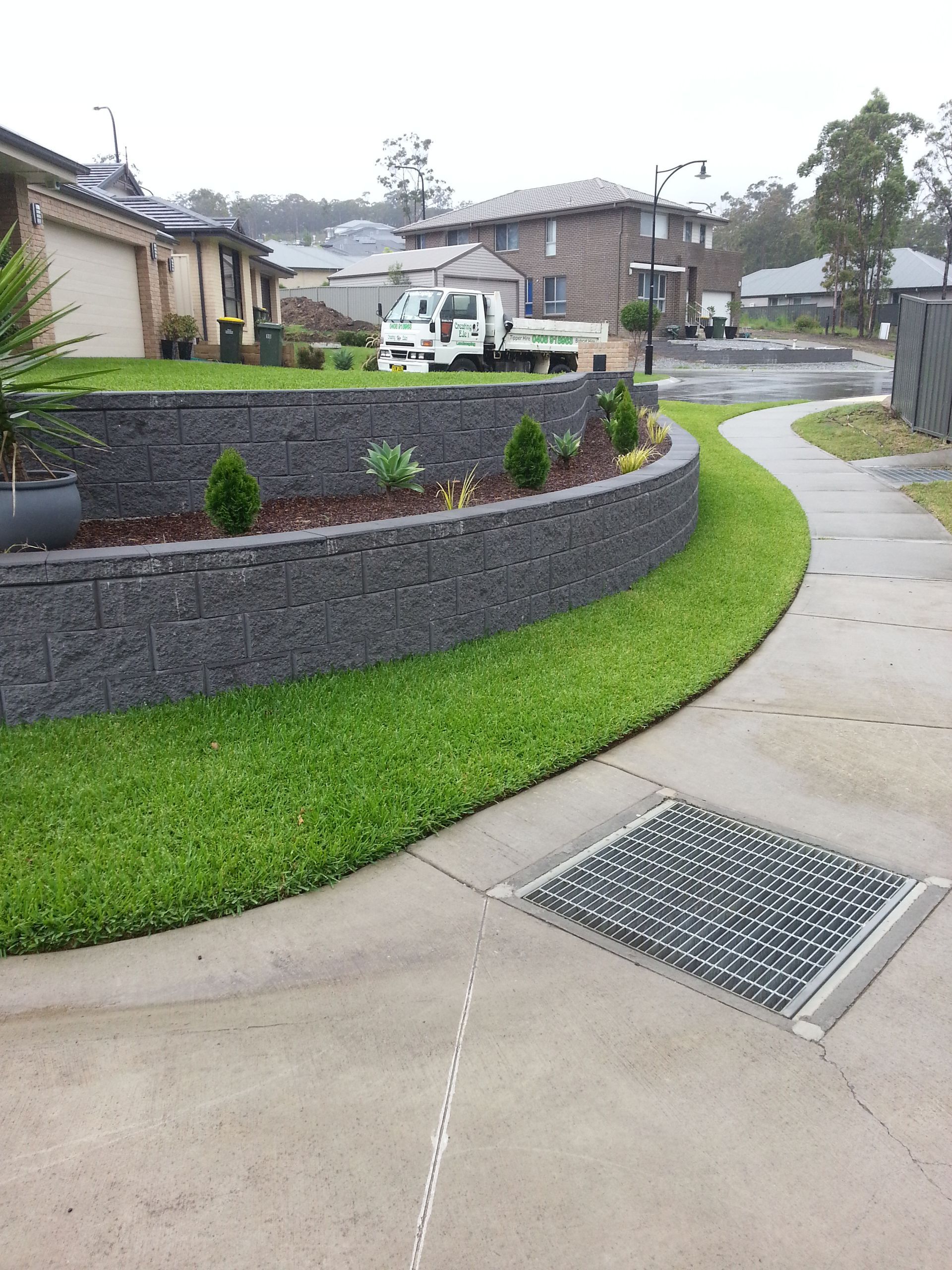 Curved retaining wall with green grass, walkway, and a storm drain in a suburban setting.