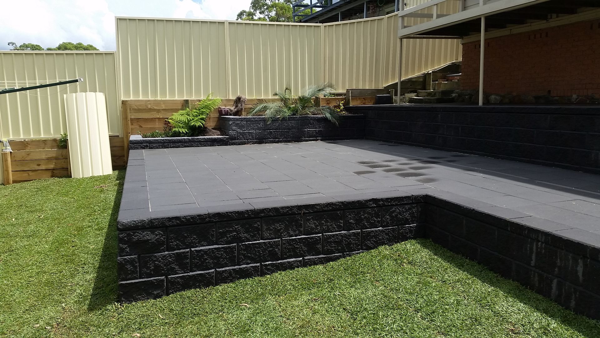 Black paved patio with retaining walls, green grass lawn, and a corrugated fence.