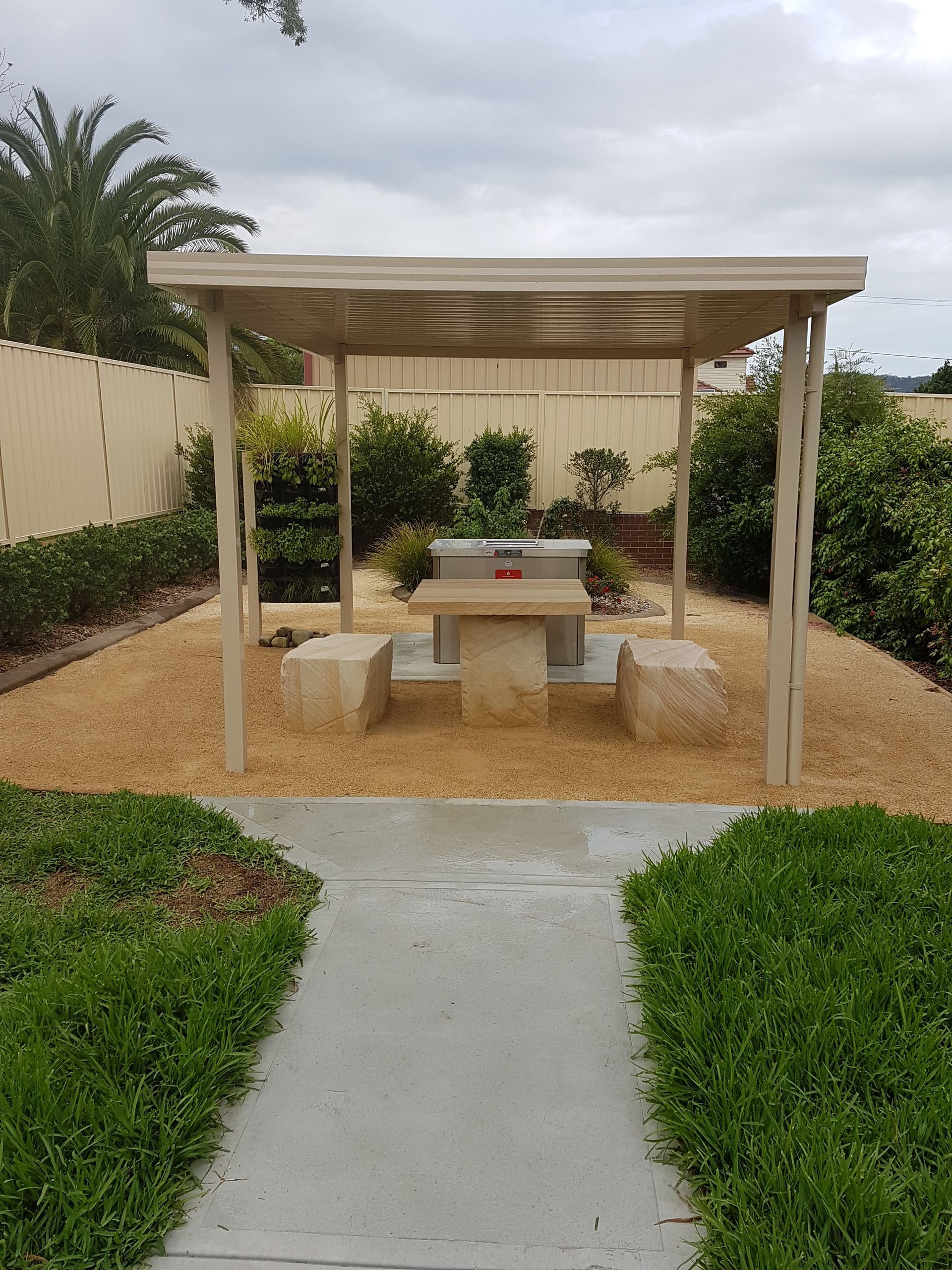 A shaded outdoor picnic area with stone table and benches, surrounded by plants and a walkway.