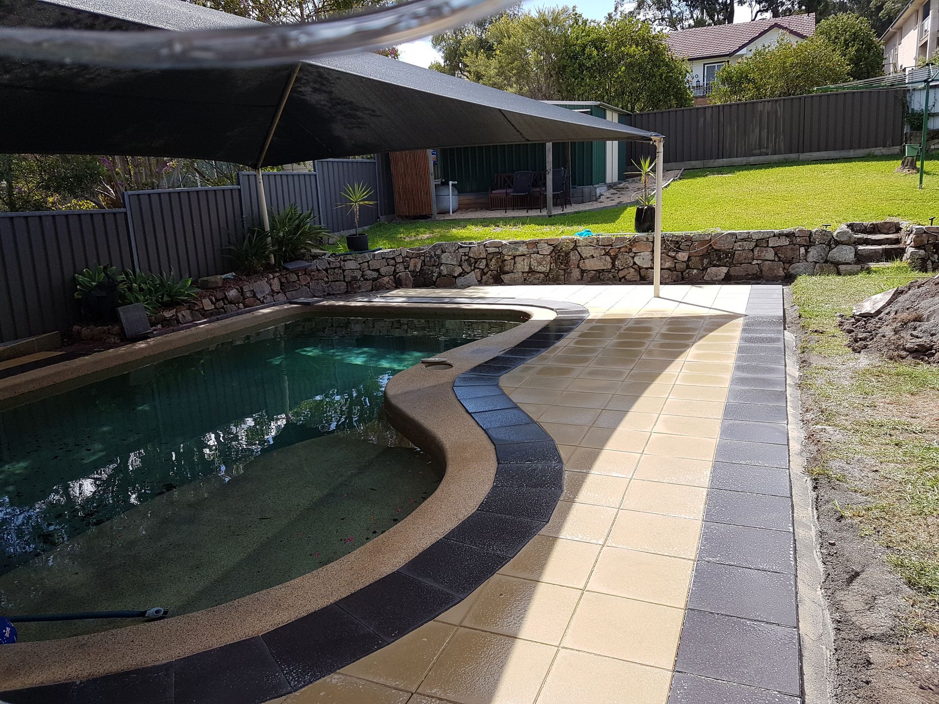 Pool with paved patio, backyard setting with shade sail and grassy lawn.