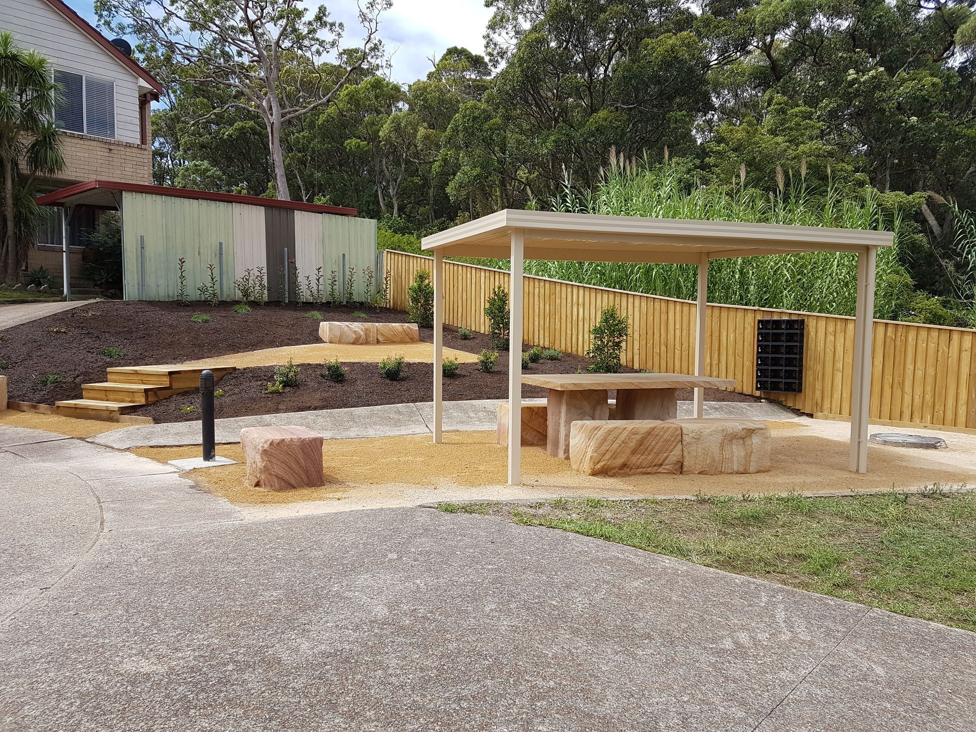 Outdoor seating area: tan pergola, stone table/seats, wooden fence, steps, shed, greenery.