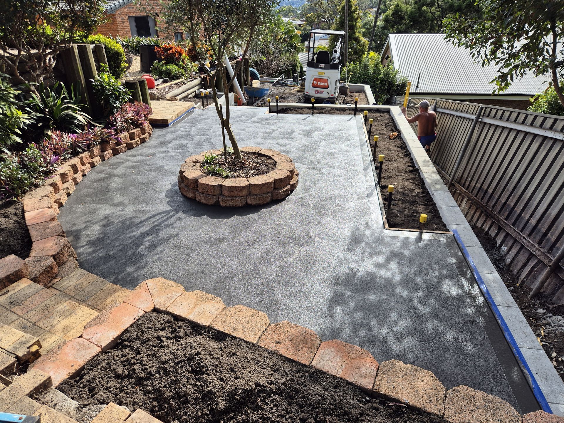 A backyard patio under construction, featuring brick borders, a small tree, and a mini excavator — Creating Eden Landscaping In Boolaroo, NSW