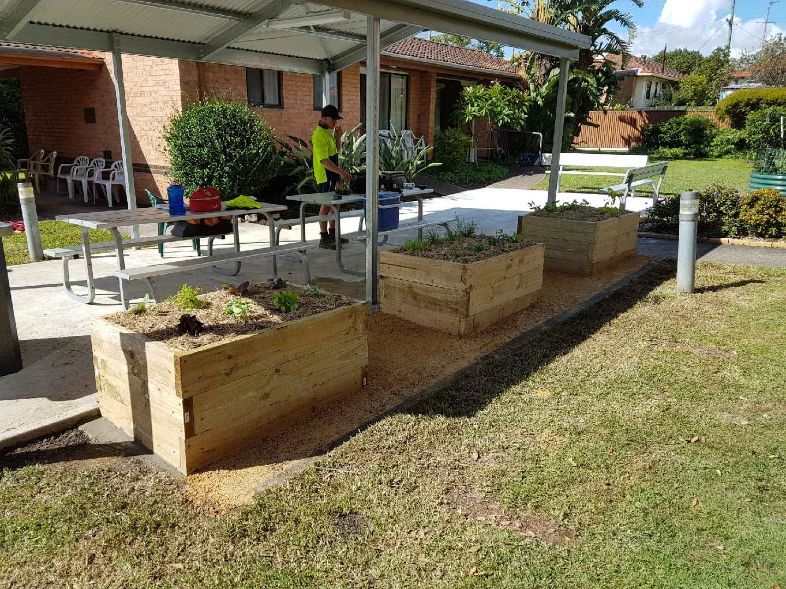 A Man Is Standing Under a Canopy Next to A Row of Wooden Planters — Creating Eden Landscaping In Speers Point, NSW