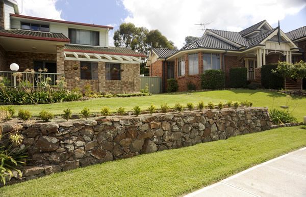 A Stone Wall Surrounds a Lush Green Lawn in Front of A House — Creating Eden Landscaping In Warners Bay, NSW