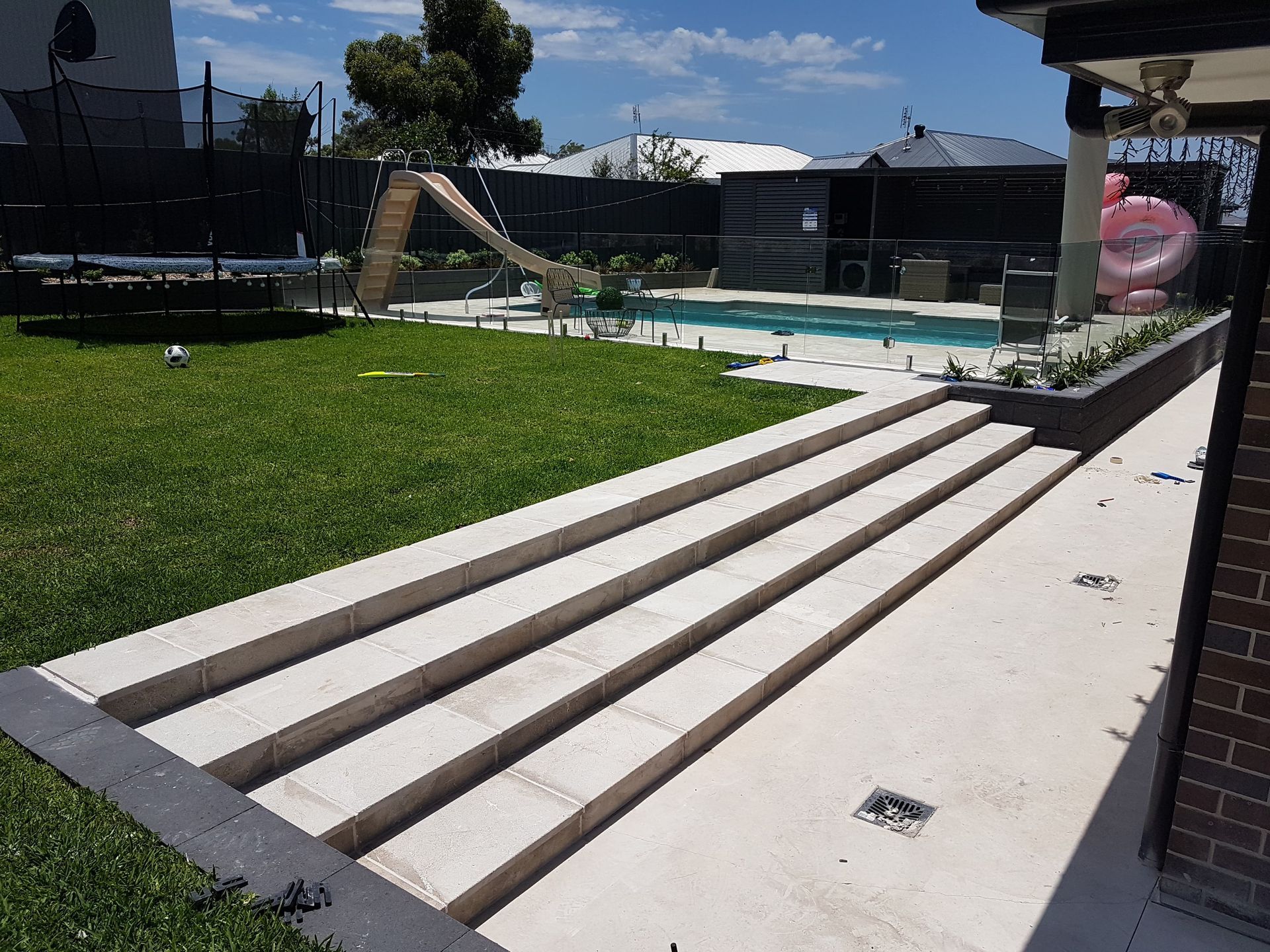 Outdoor space with steps leading to a pool, green lawn, trampoline, and a slide.