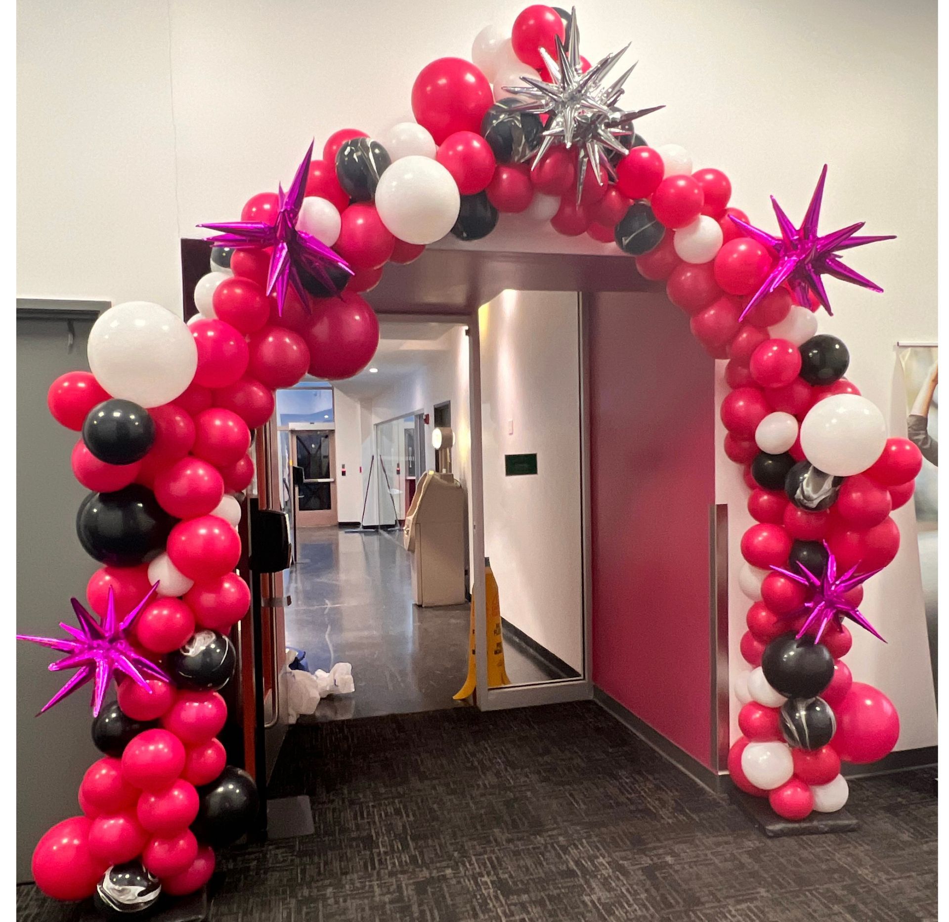 A red , white , and black balloon arch in a hallway.