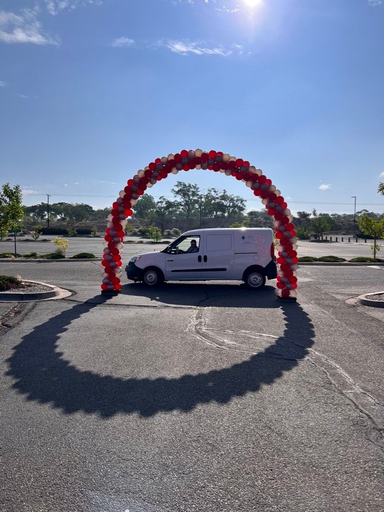 A white van is driving under a red balloon arch in a parking lot.