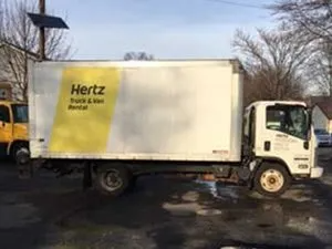 Hertz rental truck, white box truck with yellow logo, parked on a wet asphalt surface.