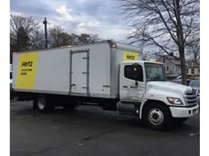 Hertz rental truck with a white cab and a white box. Yellow Hertz logo. Parked outdoors.