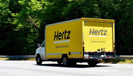 Yellow Hertz box truck on a highway, green trees in the background.