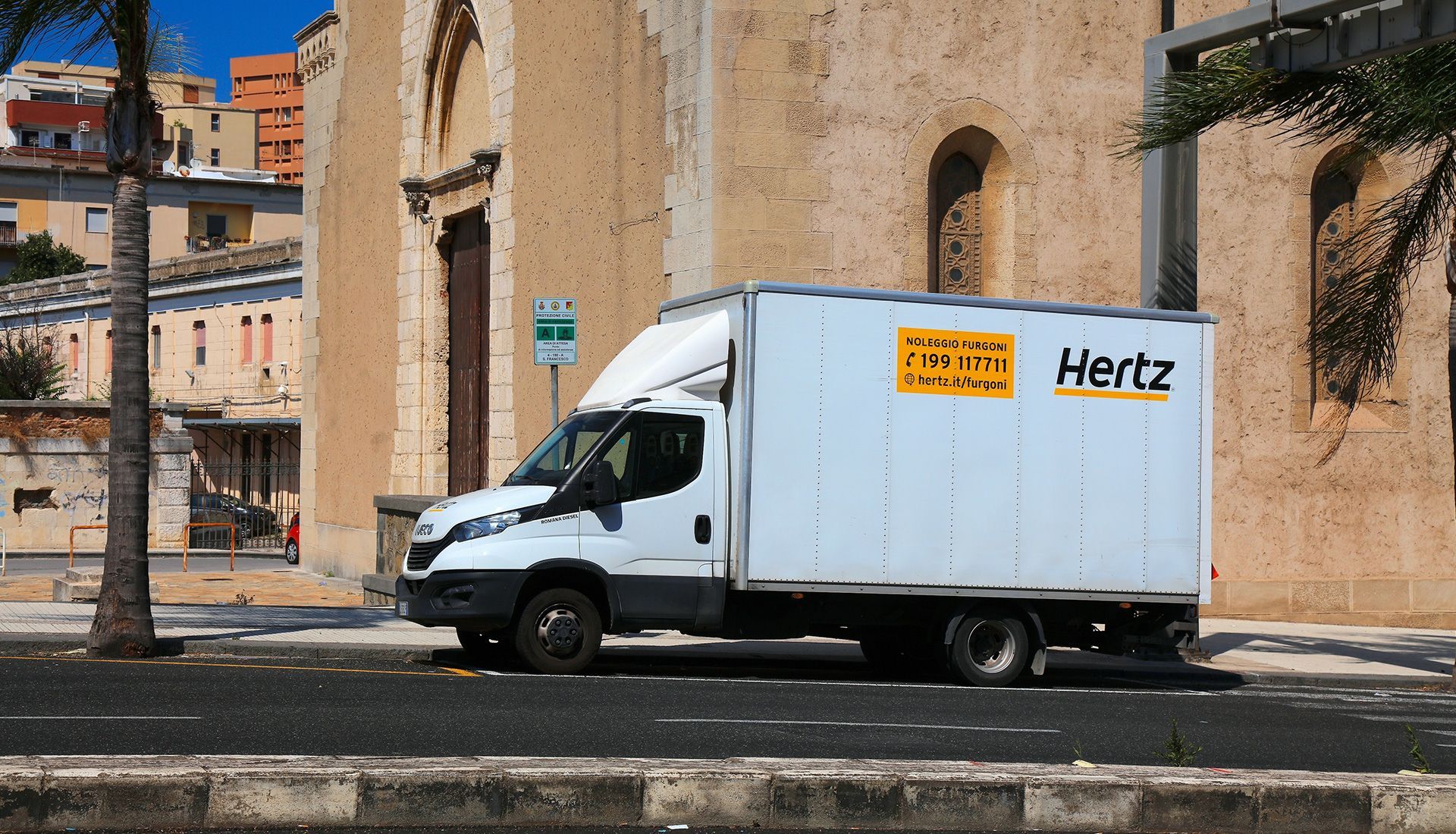 White Hertz box truck parked on a street in front of a tan building.