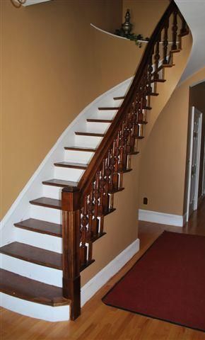 Curving wooden staircase with dark railing, white risers, and brown treads against tan walls; burgundy rug.