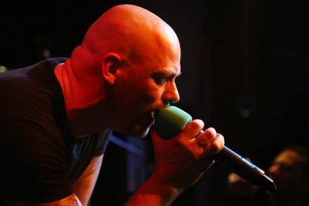Kale zanger in rood podiumlicht zingt in een microfoon op een donker podium.