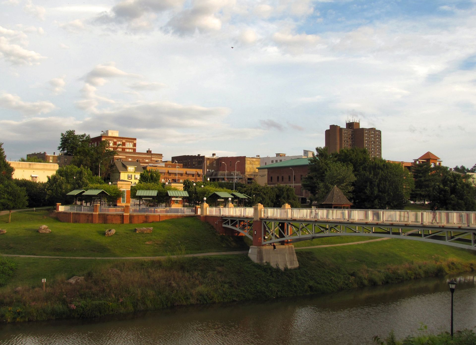 Riverfront town at sunset with a bridge, grassy bank, and buildings under a cloudy sky in Maryville, TN.