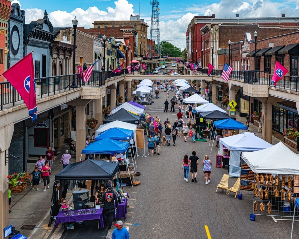 Outdoor market on a Morristown,  TN city street with tents, pedestrians, brick buildings, and pink flags under a cloudy sky. 