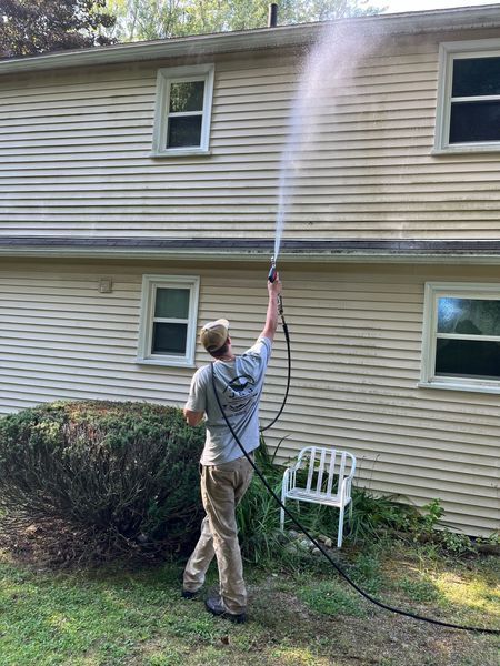 Person pressure washing the side of a beige house with windows, standing on grass near a bush.