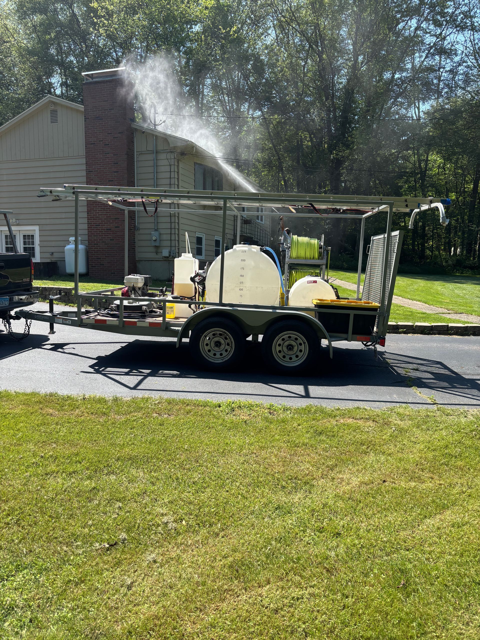 A trailer-mounted pressure washing system spraying water onto a house roof; sunny day.
