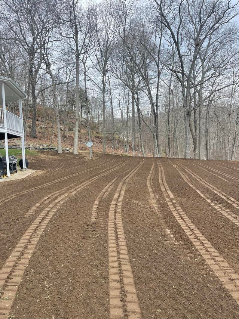 Rows of freshly tilled brown soil in a yard, leading to a treeline; a white porch is on the left.