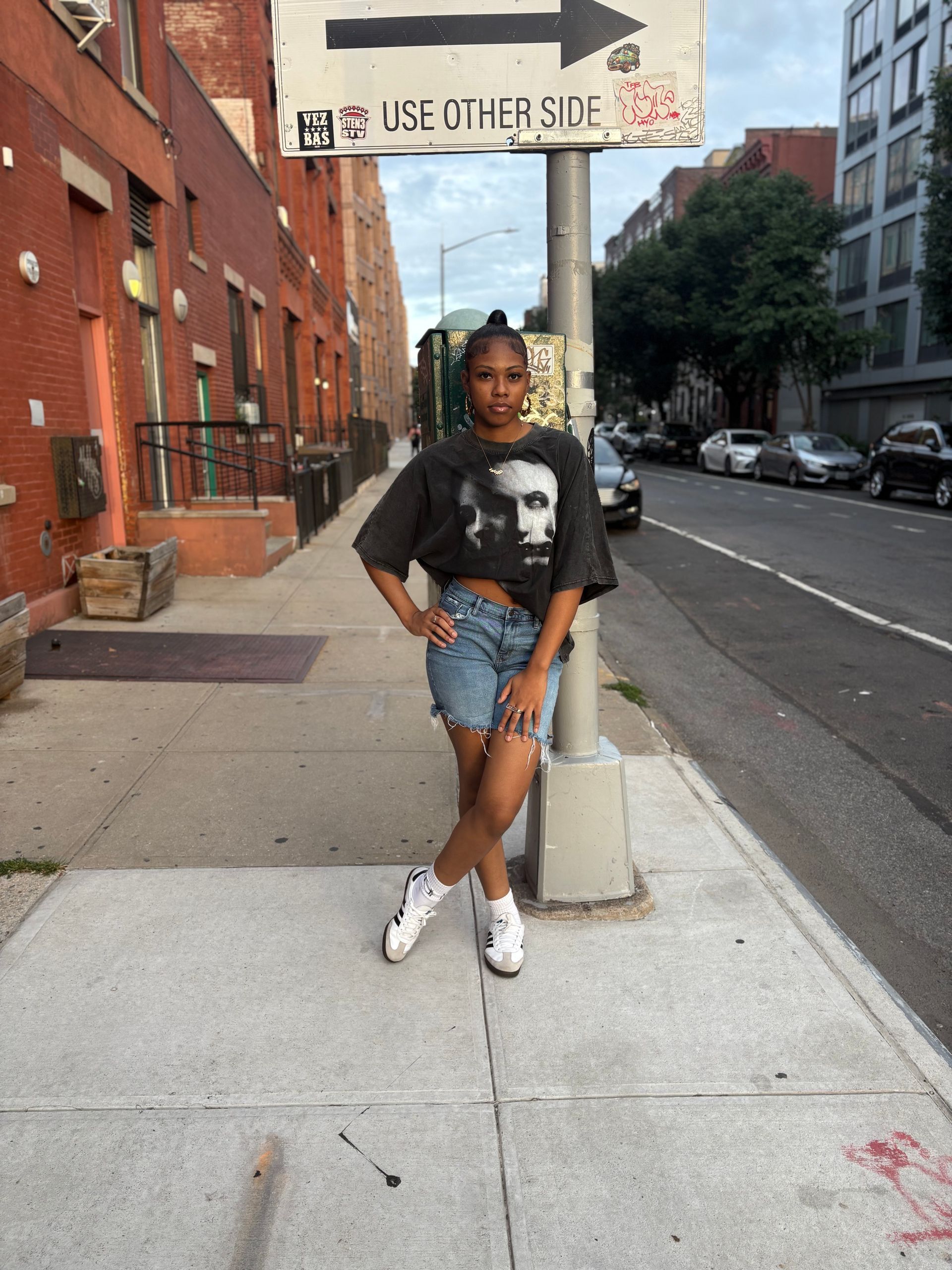 Woman in a black graphic tee and denim shorts stands on a sidewalk next to a street sign.