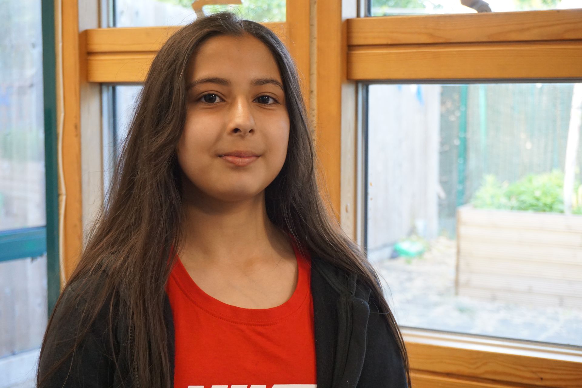 A young girl with long hair is standing in front of a window.