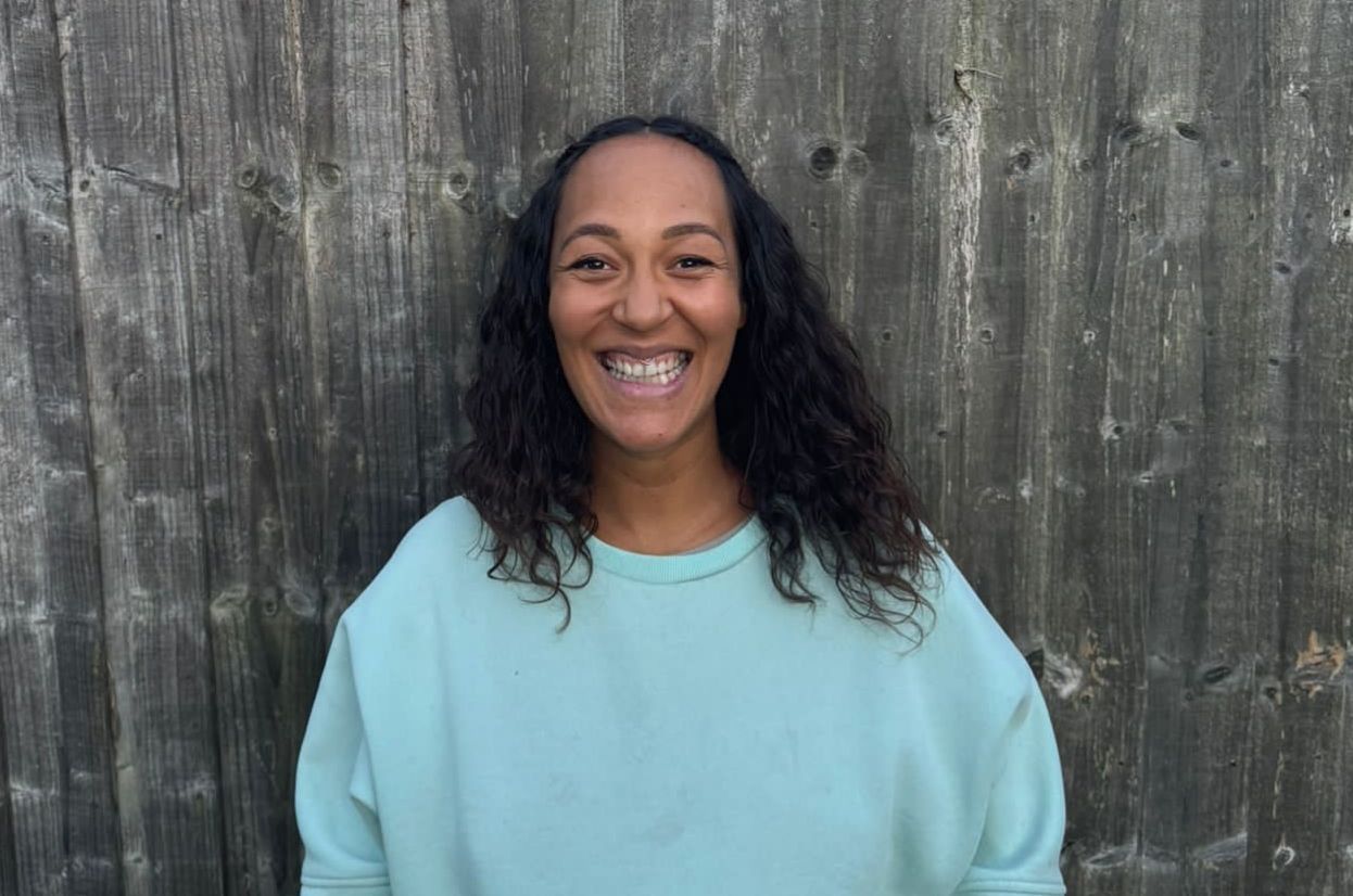 A woman in a blue sweater is smiling in front of a wooden wall.