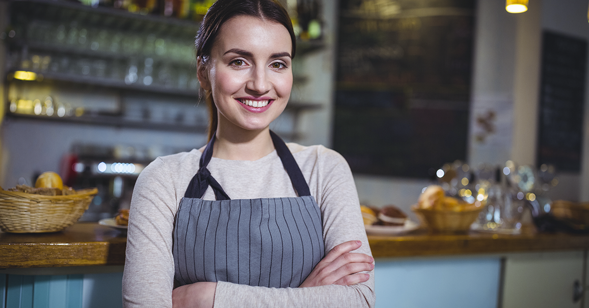 Una mujer con delantal está parada en un restaurante con los brazos cruzados.