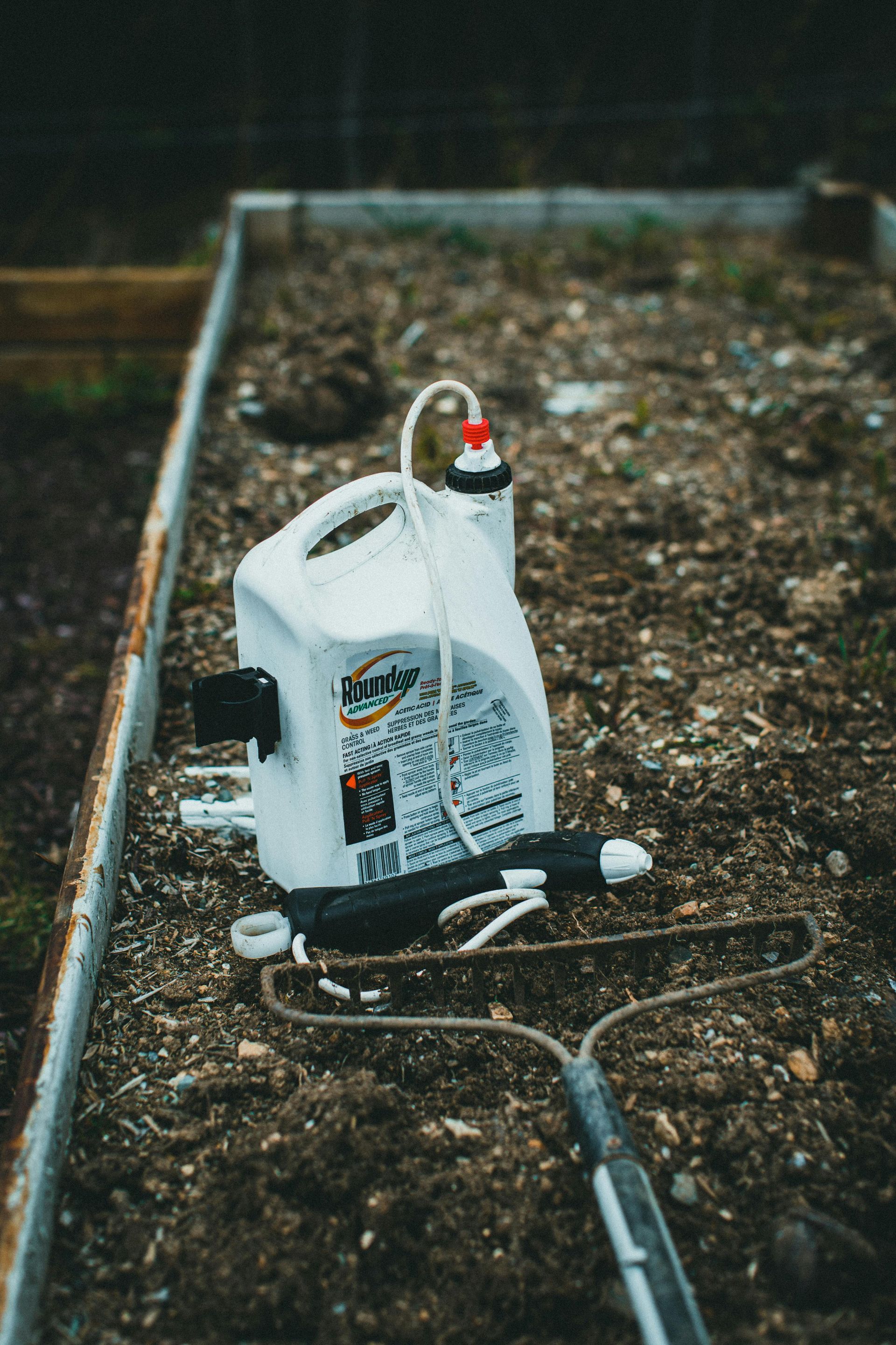 White pesticide sprayer and rake rest in a garden bed filled with soil.