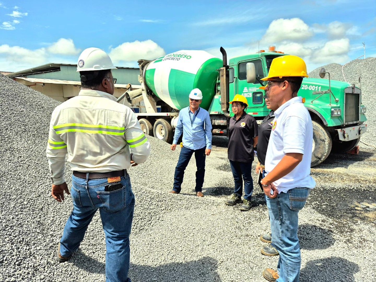 Hombres con cascos y ropa informal de pie cerca de un camión hormigonera verde y pilas de grava, posiblemente en un sitio de construcción.