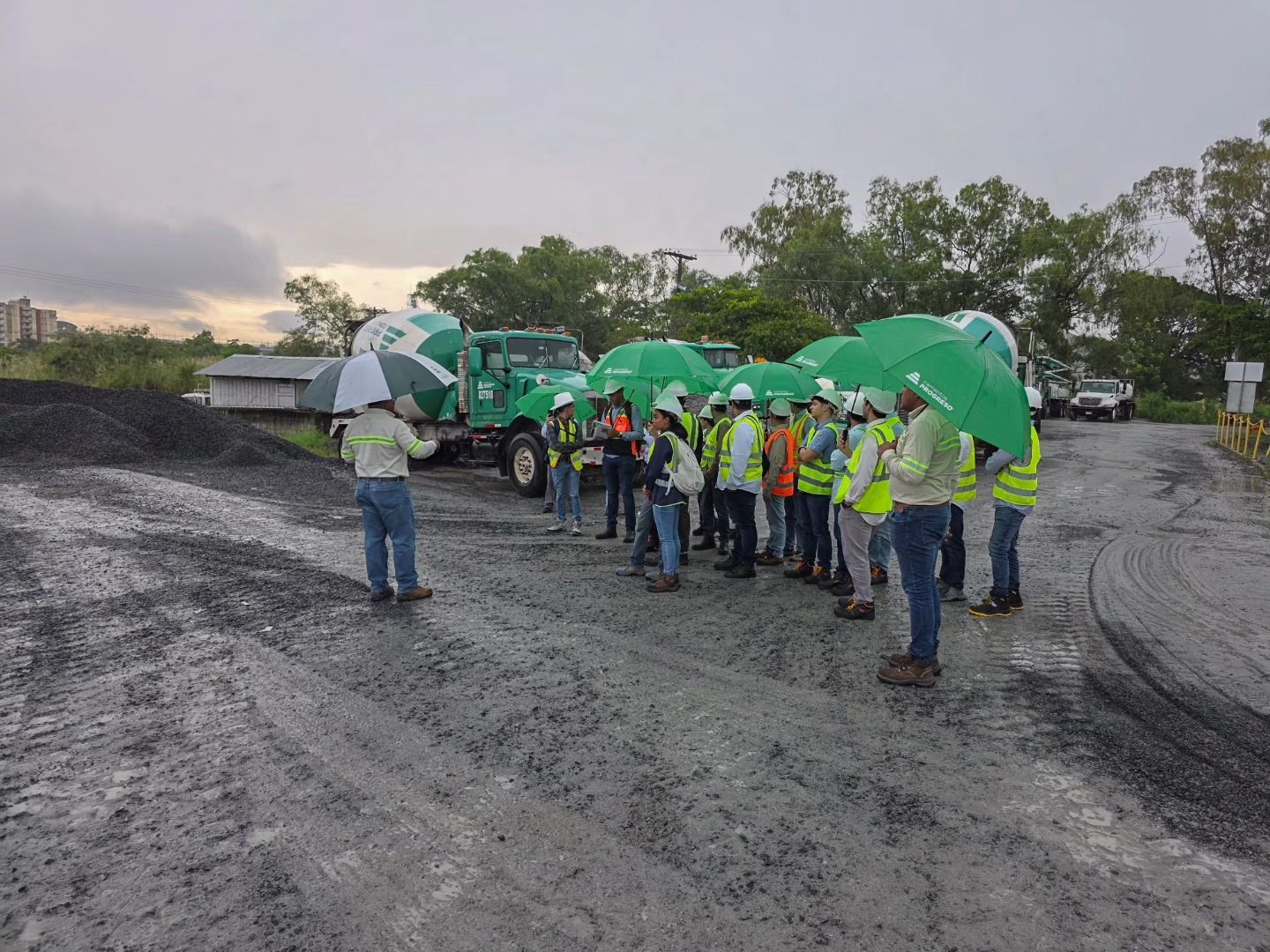 Grupo de personas con paraguas verdes, cerca de camiones, en una zona fangosa; una persona está hablando.