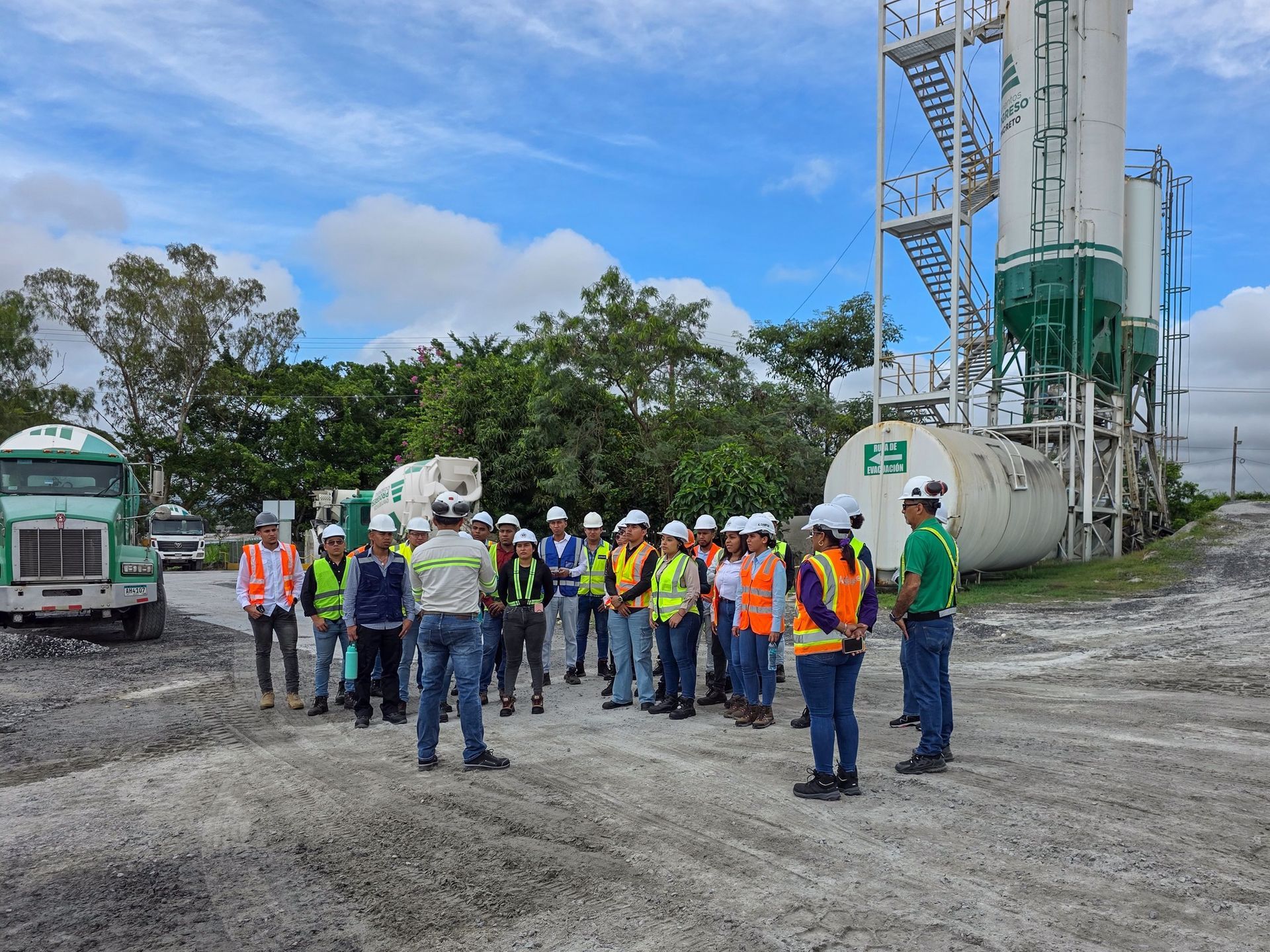 Grupo de personas con chalecos de seguridad y cascos de seguridad en un sitio industrial cerca de tanques y maquinaria.