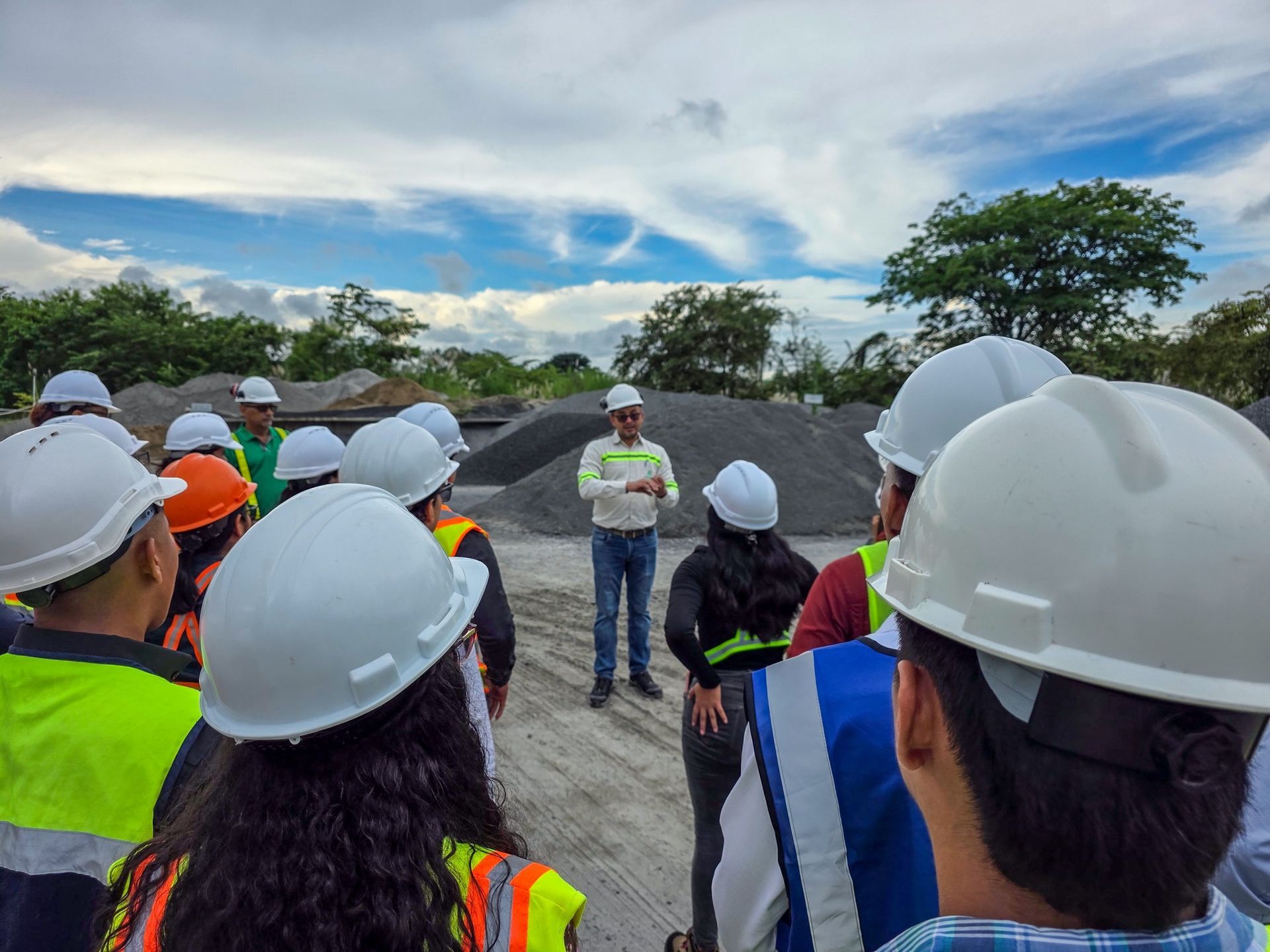 Un grupo de personas con cascos escuchan a un hombre en un sitio de construcción con montones de grava en el fondo.