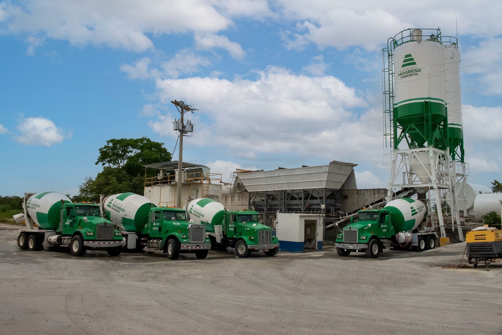 Camiones de cemento verdes estacionados en una planta de hormigón con silos y edificios bajo un cielo nublado.