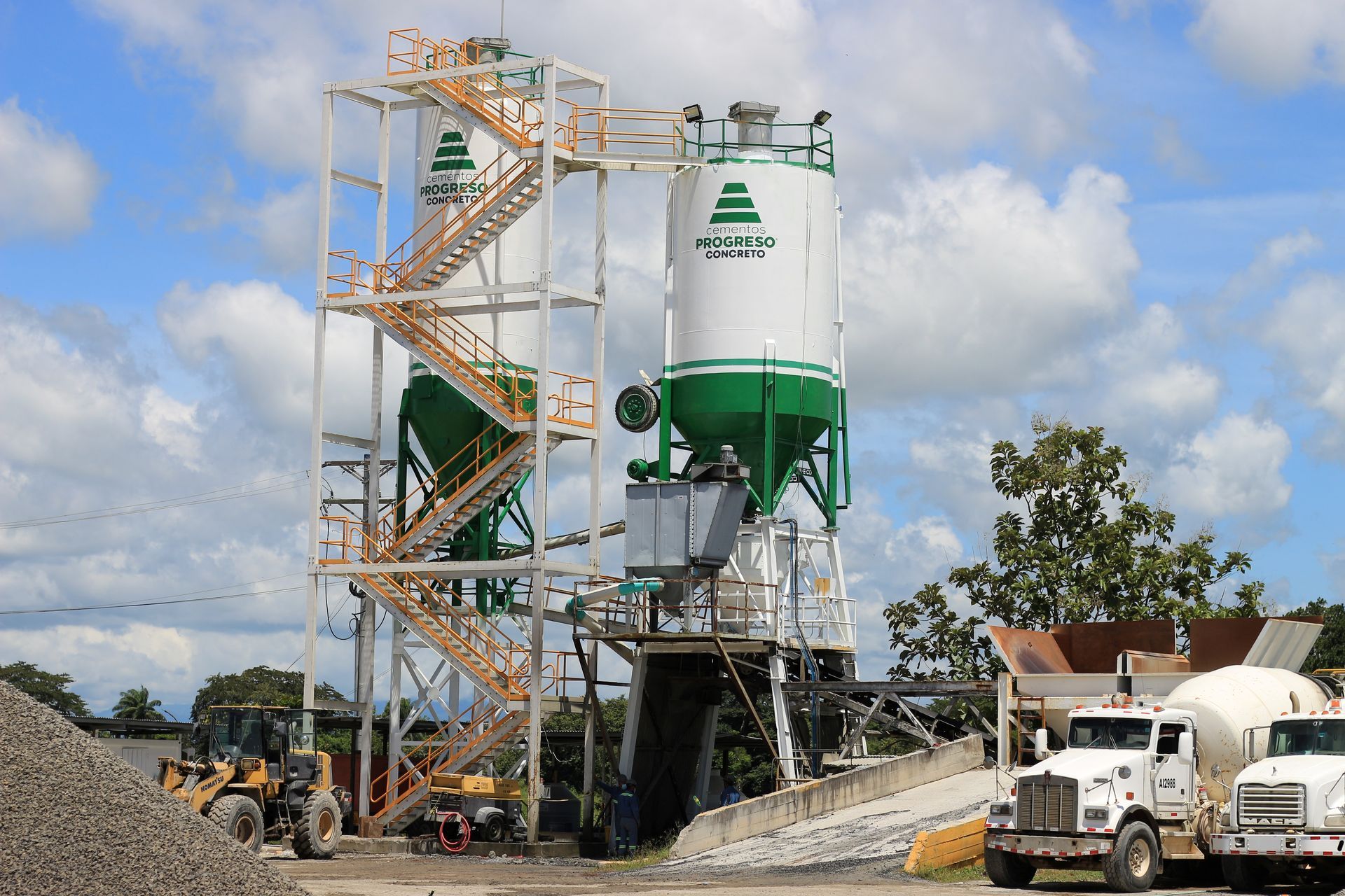 Planta de hormigón con dos grandes silos, escaleras y camiones. Combinación de colores verde y blanco.
