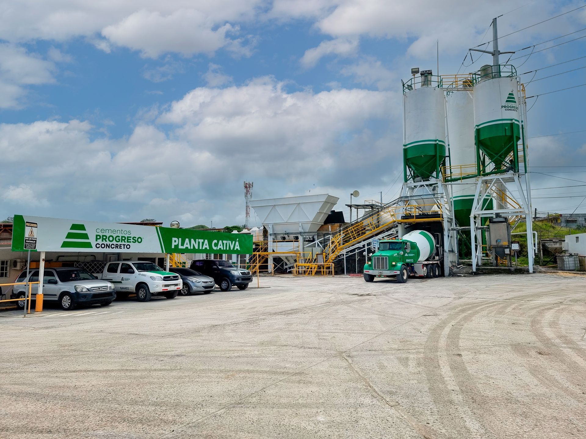 Planta de hormigón con silos, camiones y vehículos estacionados bajo un cielo parcialmente nublado.