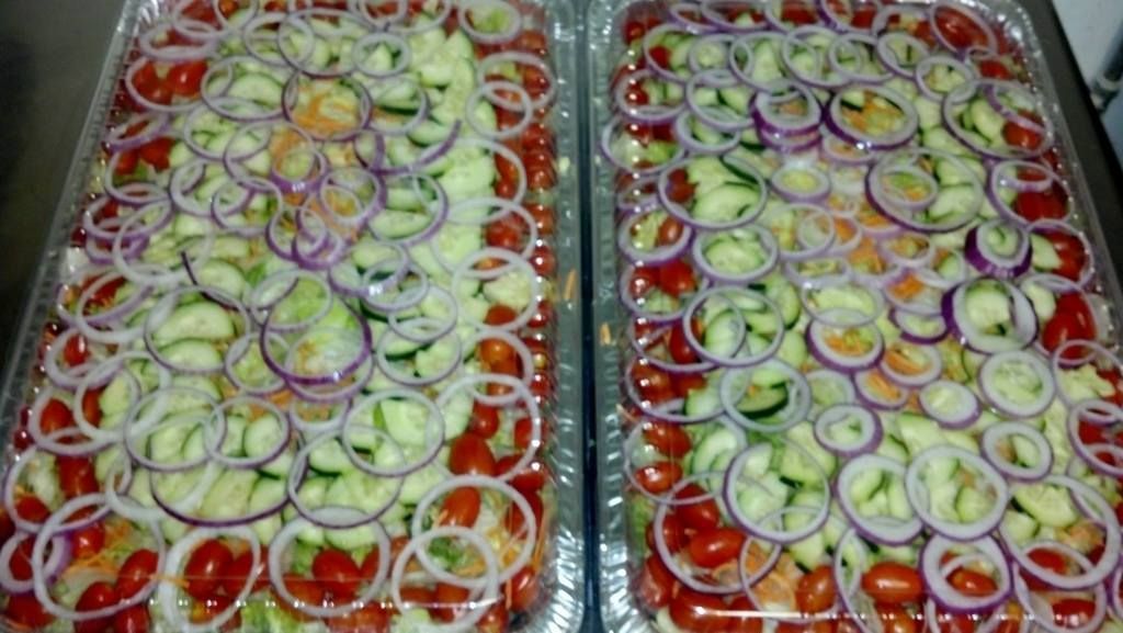 Two rectangular trays of layered sliced vegetables: tomatoes, cucumbers, and red onion.