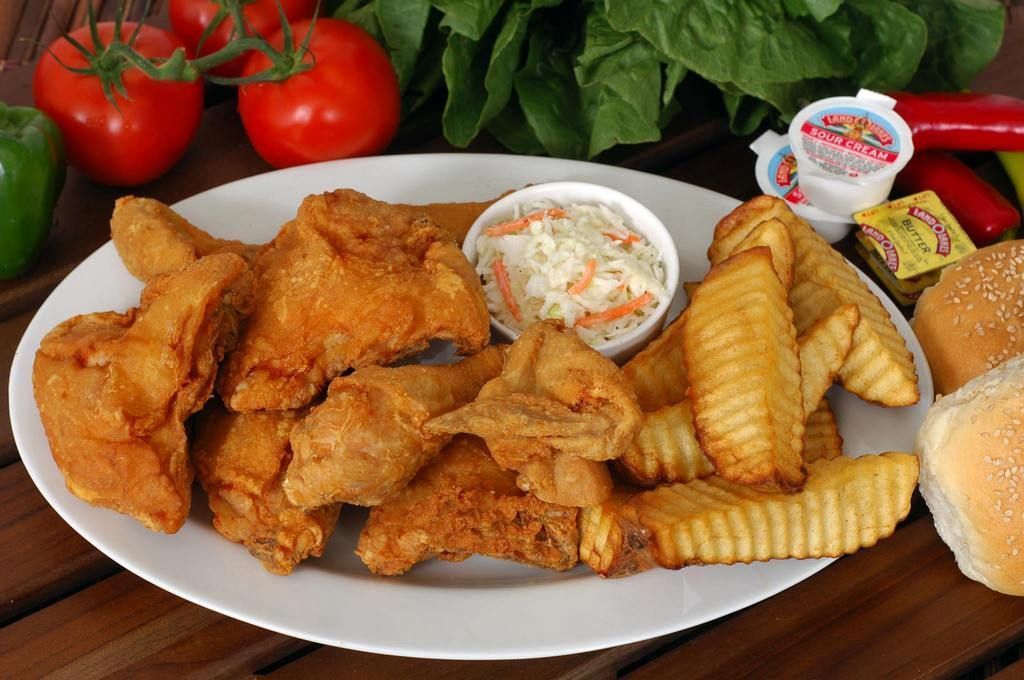 Plate of fried chicken, fries, and coleslaw with tomatoes, peppers, and bread in the background.