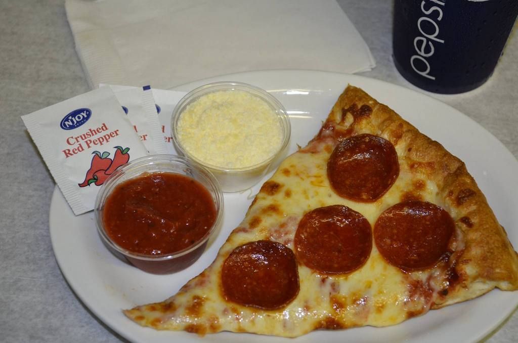 Slice of pepperoni pizza with sauce, parmesan, and crushed red pepper packets on a white plate; a Pepsi cup in the background.