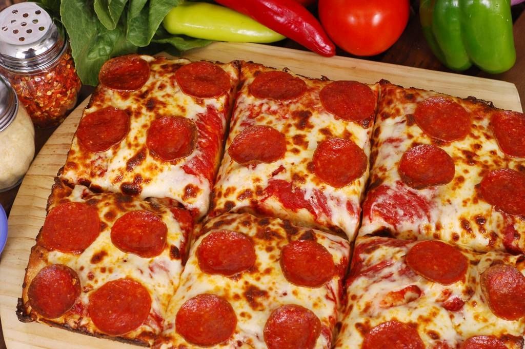 Square pepperoni pizza, cut into slices, on a wooden board with peppers and spices.