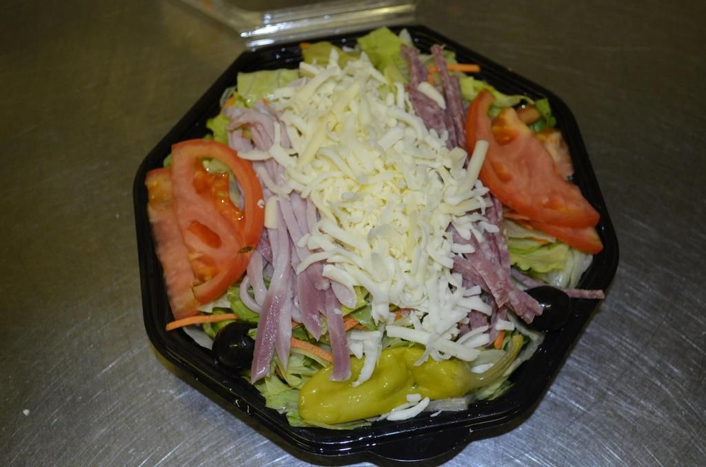 Salad in a black container with lettuce, cheese, ham, tomatoes, carrots, olives, and pepperoncini.