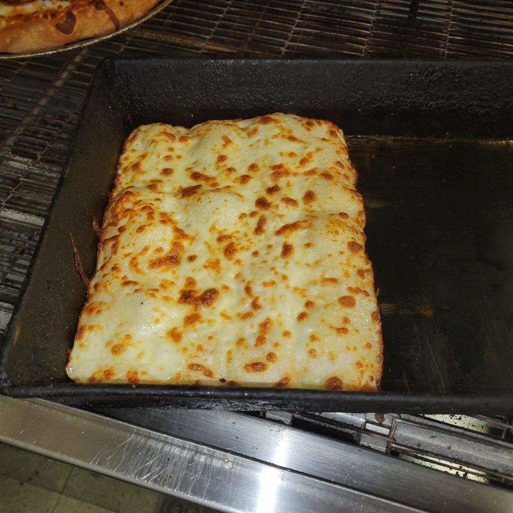 A rectangular, cheesy pizza on a dark baking sheet, with a pizza in the background.